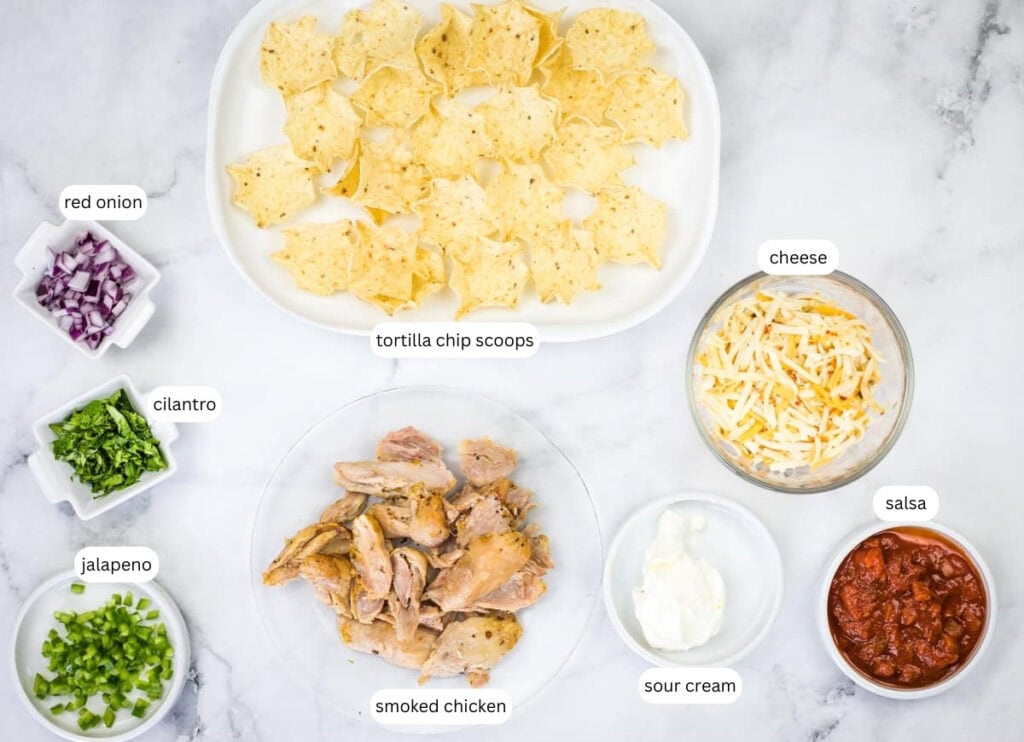 Ingredients for smoked chicken tortilla bites arranged on a white surface, including tortilla chips, smoked chicken, cheese, salsa, sour cream, jalape&ntilde;o, cilantro, and red onion.