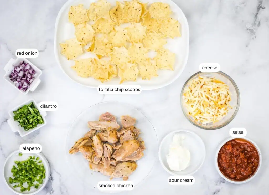 Ingredients for smoked chicken tortilla bites arranged on a white surface, including tortilla chips, smoked chicken, cheese, salsa, sour cream, jalapeño, cilantro, and red onion.