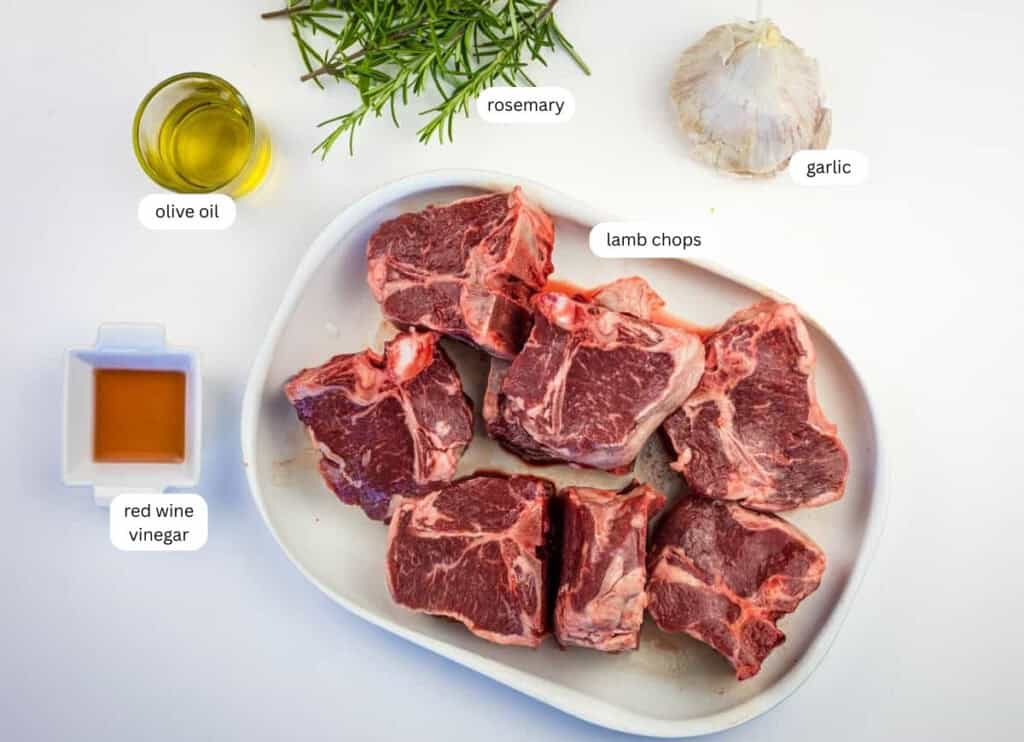 Raw lamb chops on a white plate, surrounded by labeled ingredients: olive oil, red wine vinegar, rosemary, and garlic, on a white surface.
