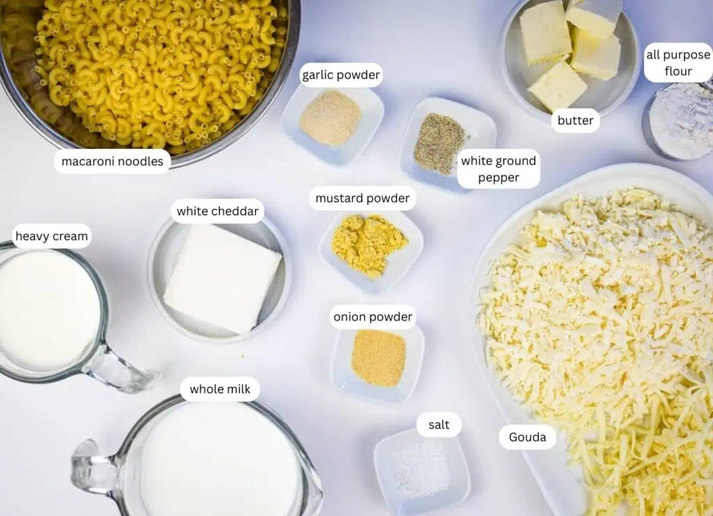 Ingredients for macaroni and cheese arranged on a white surface, including pasta, cheese, milk, cream, butter, flour, and various powders and seasonings.