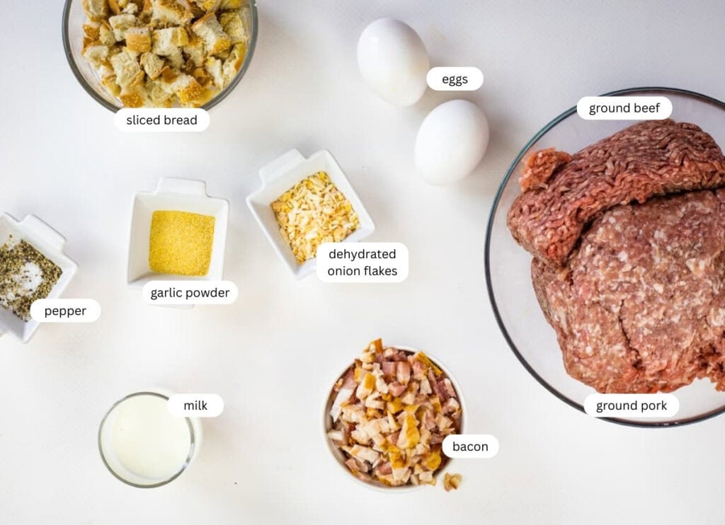 A variety of smoked meatloaf ingredients on a white surface, including ground beef, ground pork, eggs, bacon, milk, sliced bread, pepper, garlic powder, and dehydrated onion flakes.