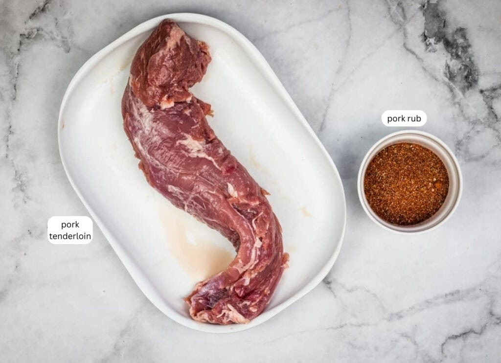 Raw pork tenderloin on a white tray next to a small bowl of pork rub seasoning, set on a marble surface.