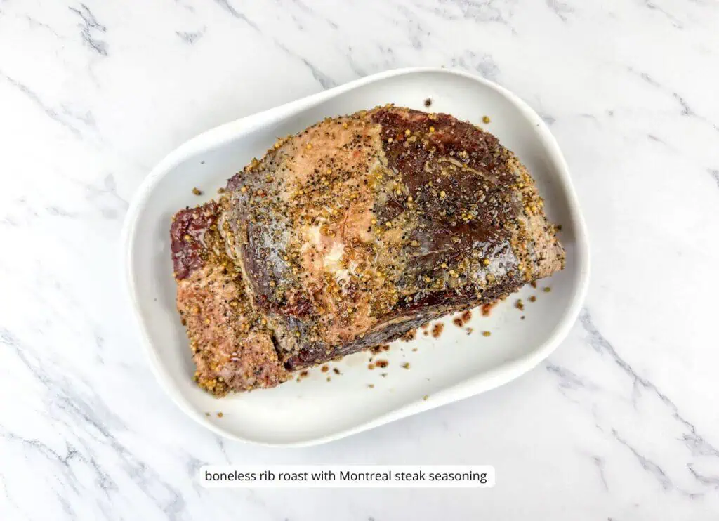 A boneless rib roast coated with Montreal steak seasoning on a white oval dish, set on a marble surface.