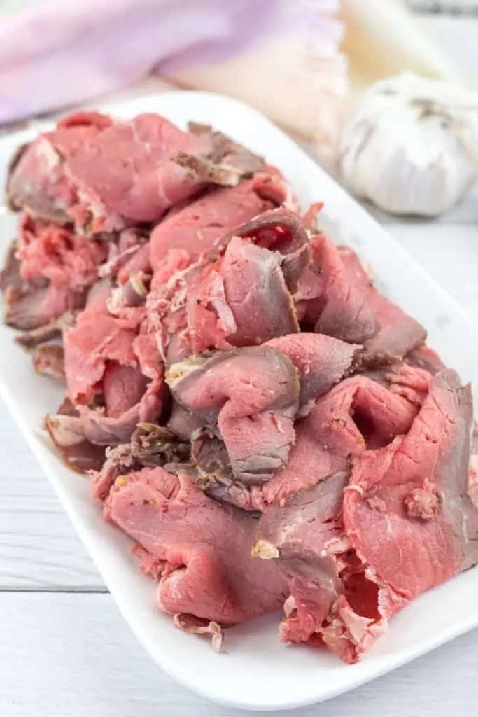 Thin slices of rare roast beef are arranged on a white rectangular plate, with a garlic bulb and a cloth in the background.