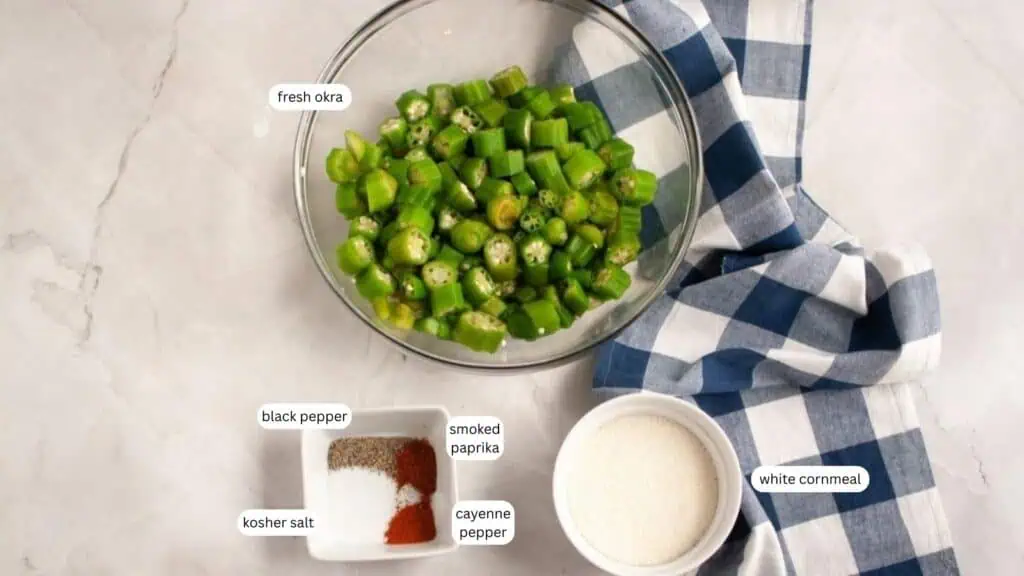A glass bowl of sliced fresh okra, a bowl of white cornmeal, spices (kosher salt, black pepper, smoked paprika, cayenne pepper), and a blue-checkered cloth on a countertop.