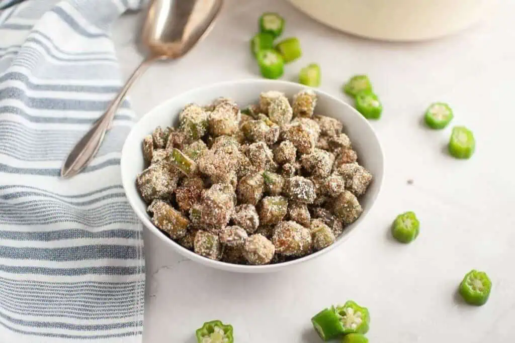 A white bowl filled with breaded and fried okra pieces, with sliced okra and a spoon nearby on a white surface.
