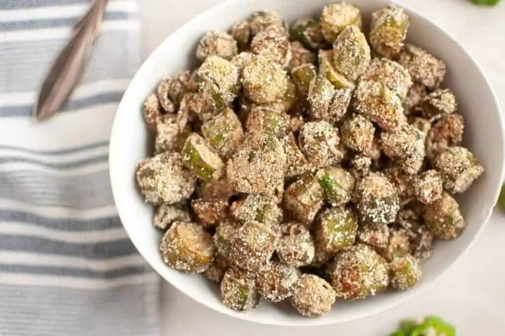 A white bowl filled with breaded and fried okra pieces on a light surface beside a striped cloth napkin.