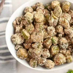 A white bowl filled with fried okra pieces on a light surface beside a striped cloth napkin.