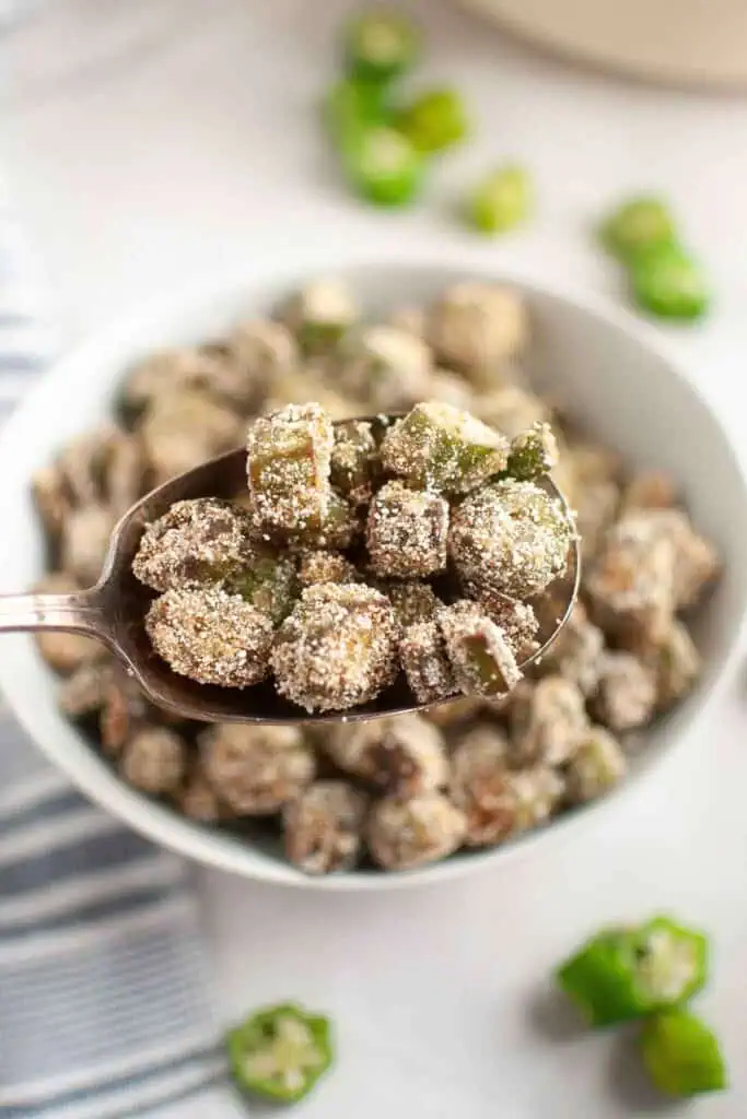 A close-up of a spoon holding fried okra coated in cornmeal, with a bowl of more fried okra and sliced okra pieces in the background.