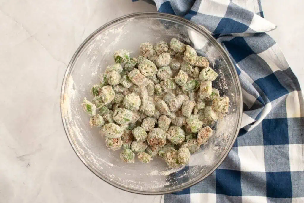 A glass bowl filled with sliced okra coated in flour on a white surface next to a blue and white checkered cloth.