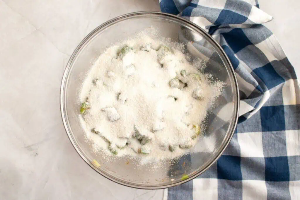 A glass bowl containing sliced green vegetables covered with a layer of granulated sugar, next to a blue and white checkered cloth on a marble surface.