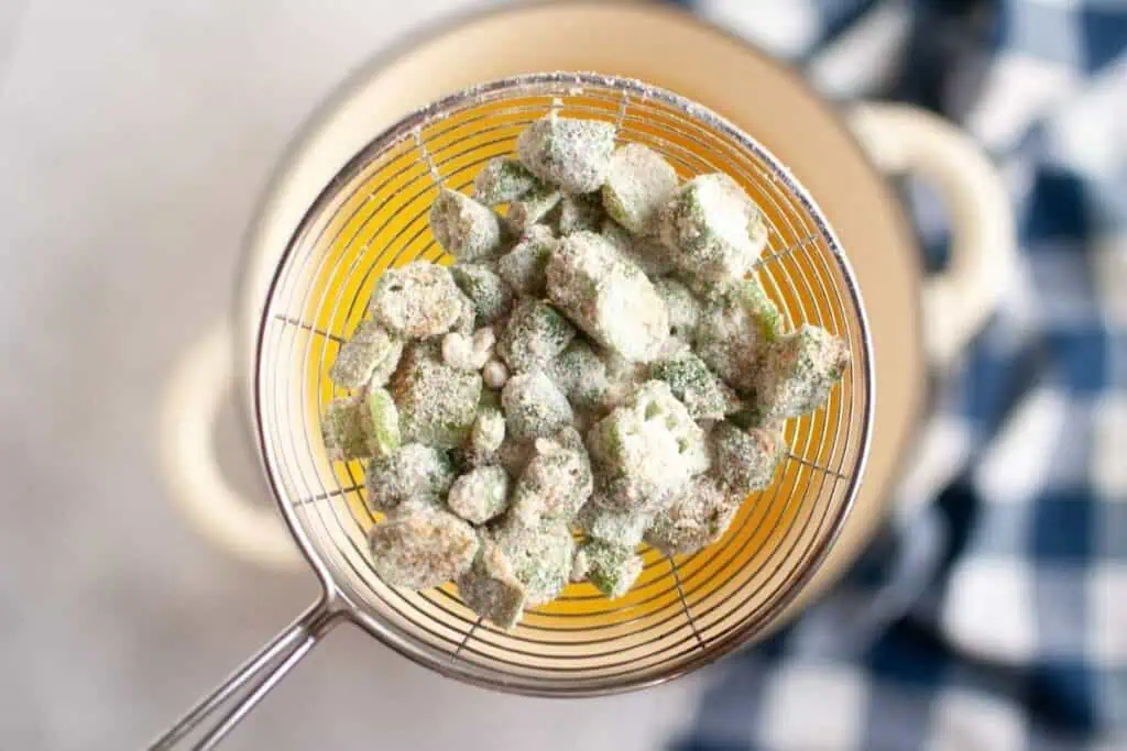 A metal strainer holds battered okra pieces over a pot, with a blue and white checkered cloth in the background.