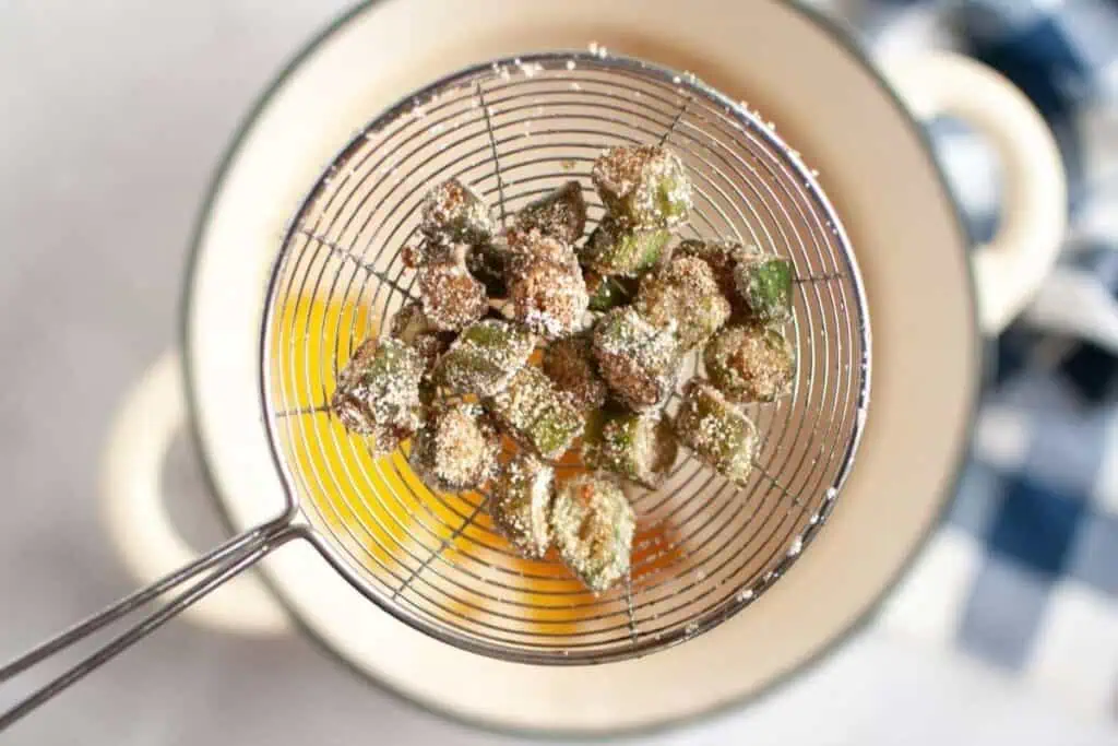 A metal strainer holds pieces of breaded and fried okra above a pot of oil.