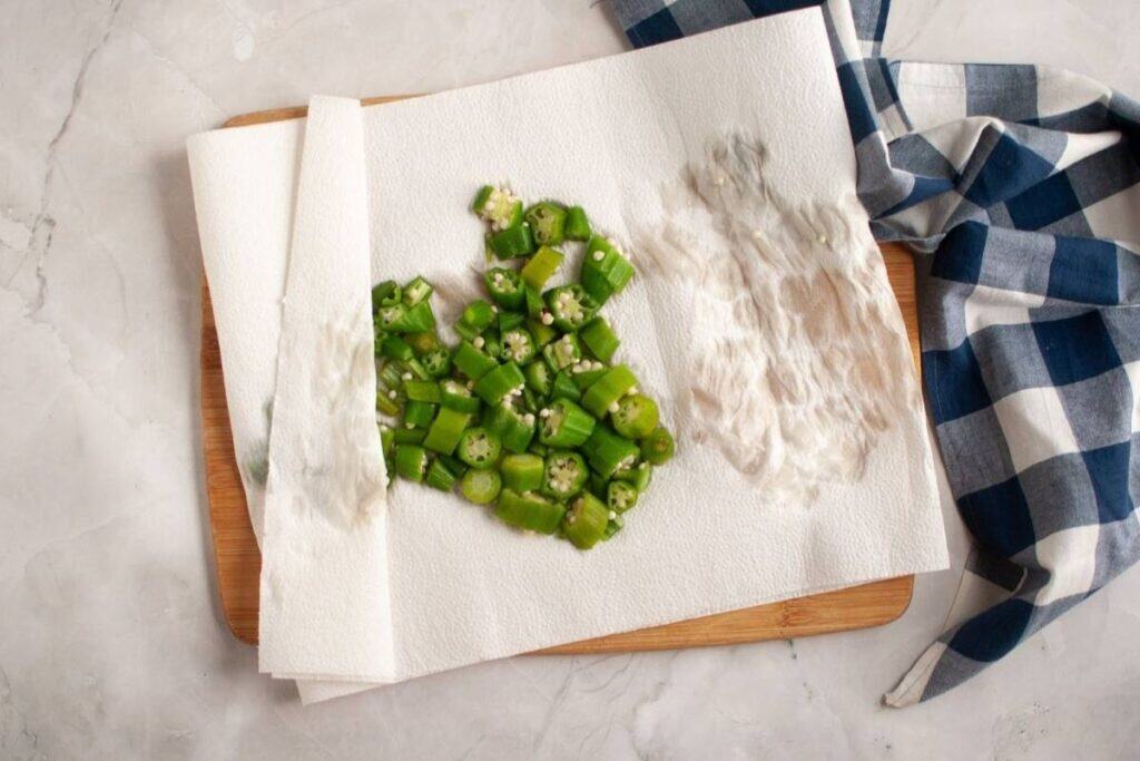 Chopped okra pieces on a paper towel-lined wooden board next to a blue and white checkered cloth on a marble surface.