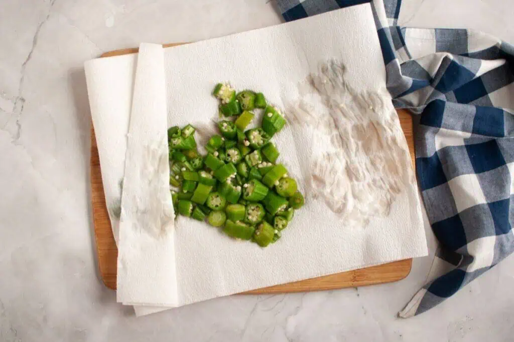 Chopped okra pieces on a paper towel-lined wooden board next to a blue and white checkered cloth on a marble surface.