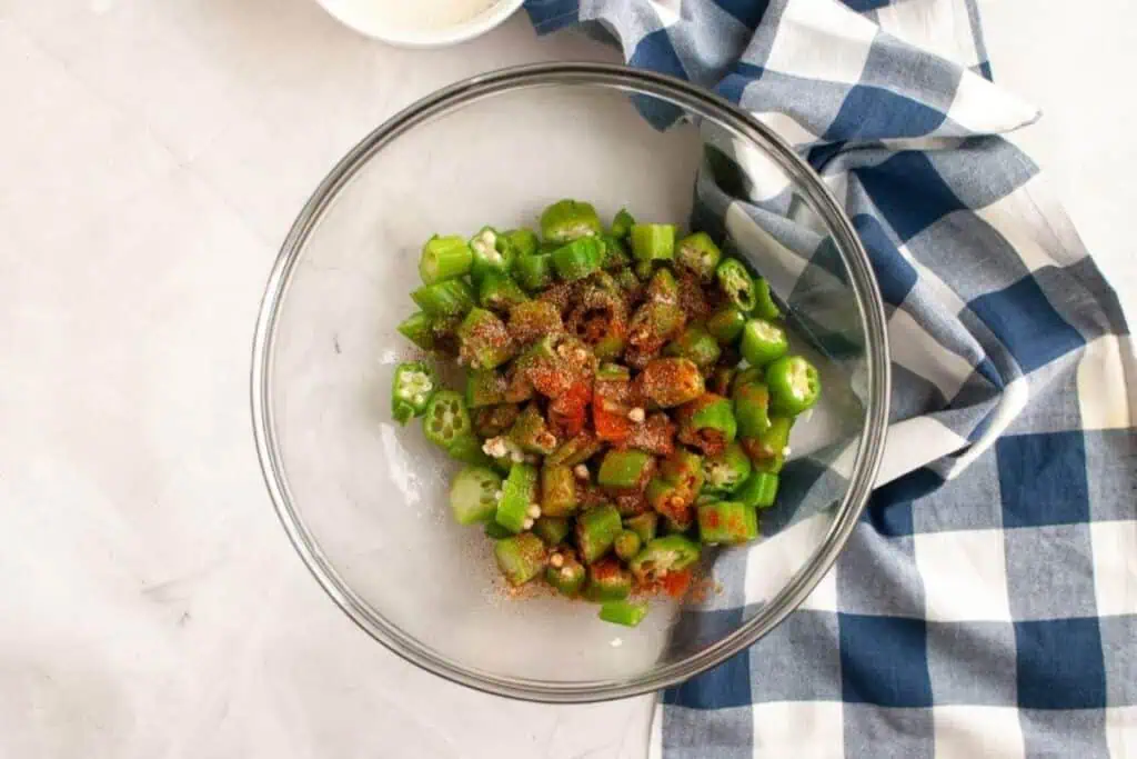 A glass bowl with sliced okra seasoned with spices sits on a light countertop next to a blue and white checkered cloth.