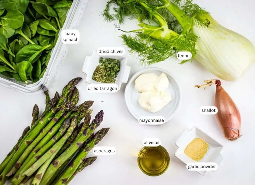 Fresh baby spinach, fennel, asparagus, and shallot are arranged on a white surface alongside small bowls of mayonnaise, olive oil, garlic powder, dried tarragon, and dried chives.