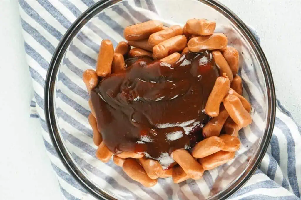 A glass bowl filled with mini sausages and topped with barbecue sauce, placed on a blue and white striped cloth.