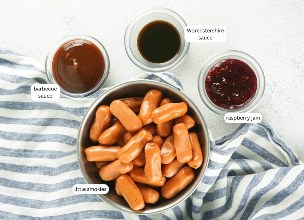 A bowl of cocktail sausages on a striped cloth, surrounded by small bowls labeled barbecue sauce, Worcestershire sauce, and raspberry jam.