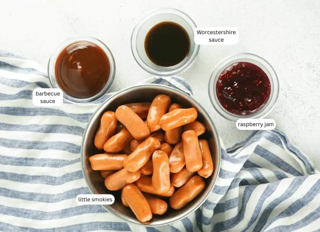 A bowl of cocktail sausages on a striped cloth, surrounded by small bowls labeled barbecue sauce, Worcestershire sauce, and raspberry jam.