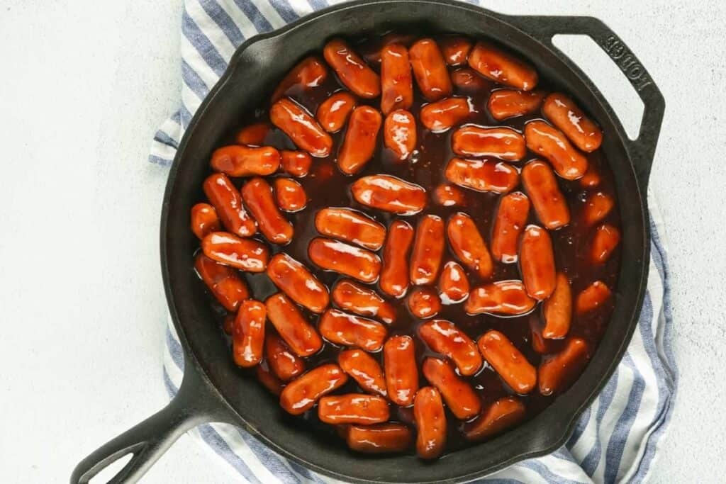 A cast iron skillet filled with cocktail sausages covered in a thick barbecue sauce, placed on a blue and white striped cloth.