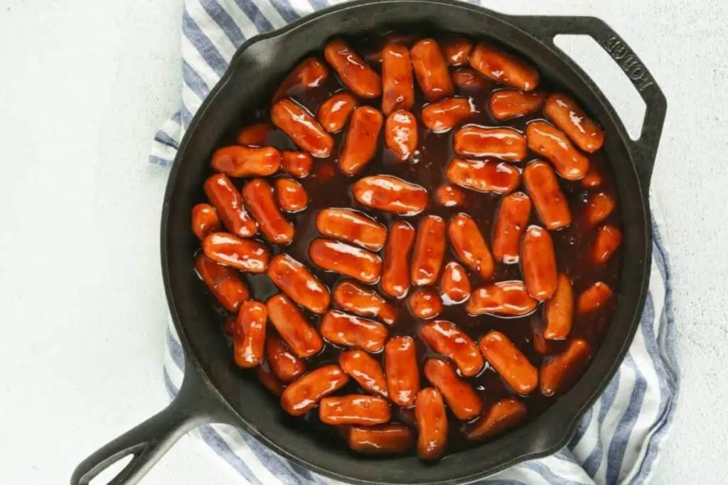 A cast iron skillet filled with cocktail sausages covered in a thick barbecue sauce, placed on a blue and white striped cloth.