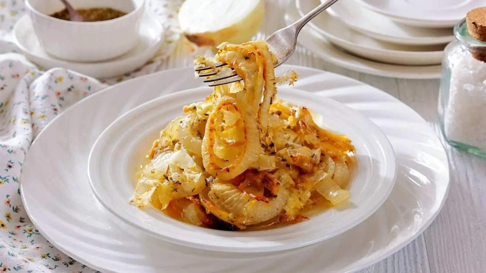 A plate of Tennessee onions garnished with herbs, with a fork lifting a portion. Plates, a jar of salt, and a bowl of seasoning are in the background.