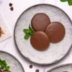 Three round chocolate-covered cookies are arranged on a parchment-lined plate with a sprig of mint, surrounded by chocolate chips and baking tools on a white surface.