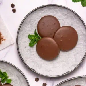 Three round chocolate-covered cookies are arranged on a parchment-lined plate with a sprig of mint, surrounded by chocolate chips and baking tools on a white surface.