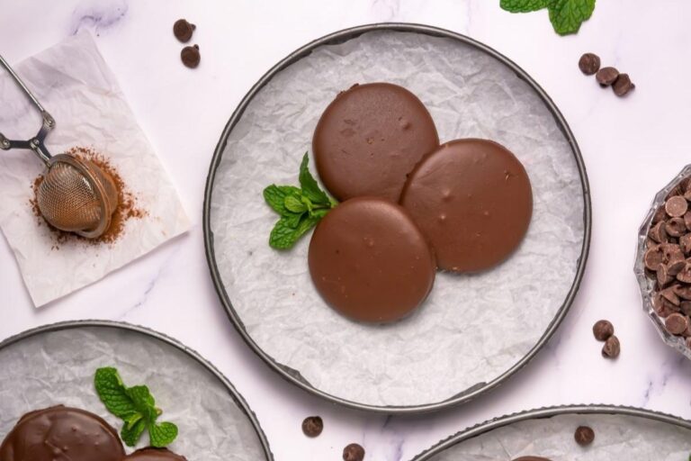 Three round chocolate-covered cookies are arranged on a parchment-lined plate with a sprig of mint, surrounded by chocolate chips and baking tools on a white surface.