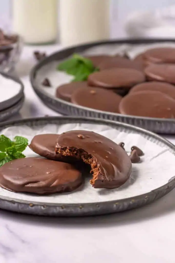 A plate of chocolate-covered cookies, one with a bite taken out, sits in the foreground with more cookies and a sprig of mint in the background.