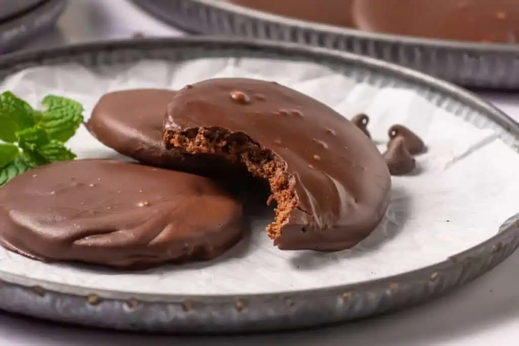 Three chocolate-covered cookies are arranged on a metal plate, with one cookie showing a visible bite. A sprig of mint is placed on the side.