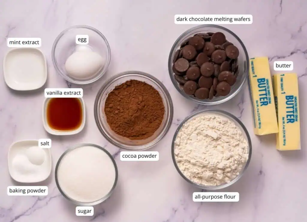 Ingredients for baking are arranged on a marble surface: butter, flour, cocoa powder, sugar, baking powder, salt, vanilla extract, mint extract, an egg, and dark chocolate melting wafers.