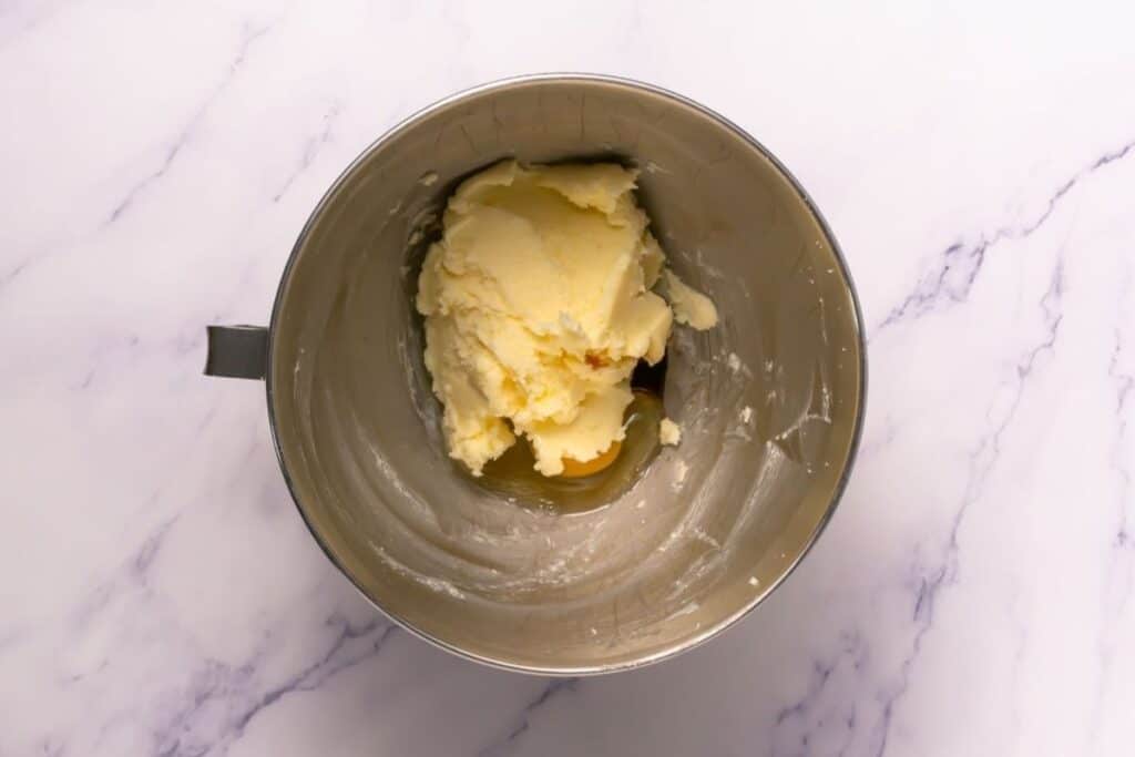 A metal mixing bowl containing creamed butter and sugar sits on a white marble surface.