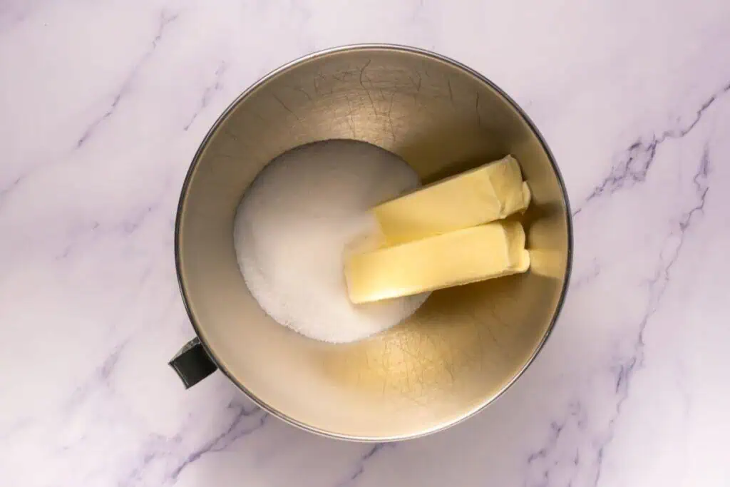 A metal mixing bowl with two sticks of butter and a pile of granulated sugar on a white marble surface.
