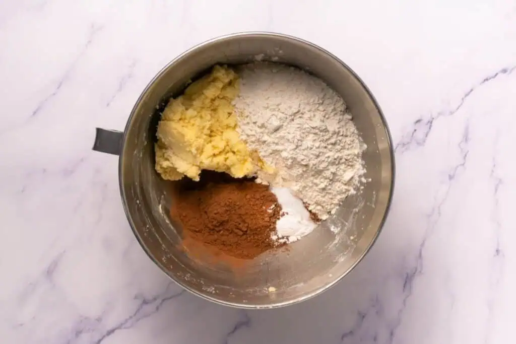 A metal mixing bowl containing butter, flour, cocoa powder, and baking powder on a white marble surface.