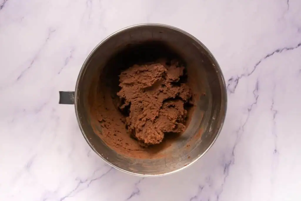 A metal mixing bowl containing a mound of chocolate cookie dough sits on a light marble surface.