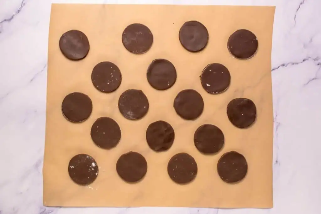 Eighteen round chocolate-covered cookies are arranged in a grid pattern on a sheet of parchment paper.