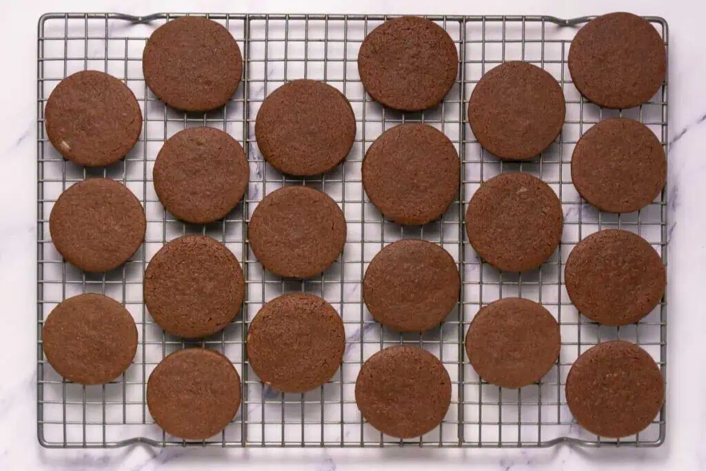 Twenty-four round chocolate cookies are arranged in rows on a wire cooling rack placed on a white surface.
