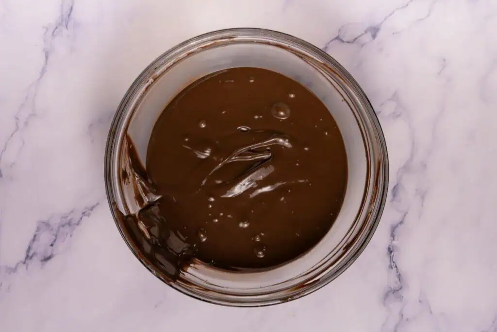 A glass bowl filled with melted chocolate sits on a white marble surface.