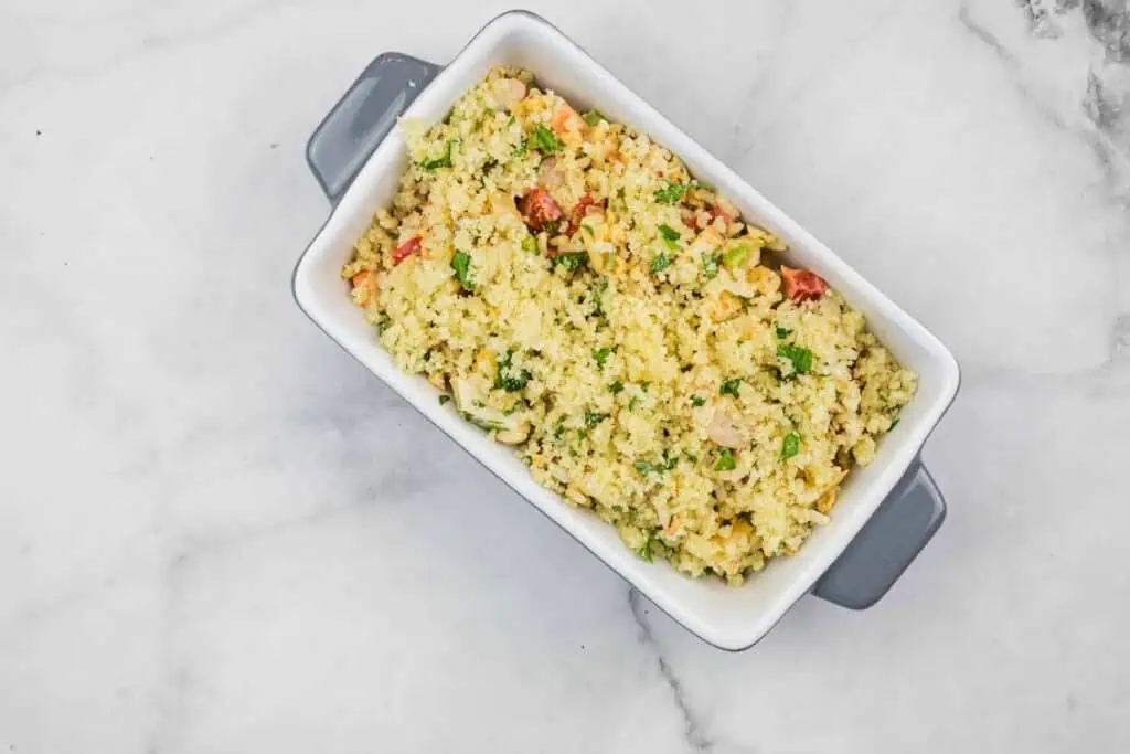 Rectangular white baking dish filled with cooked couscous mixed with chopped vegetables and herbs on a marble surface.