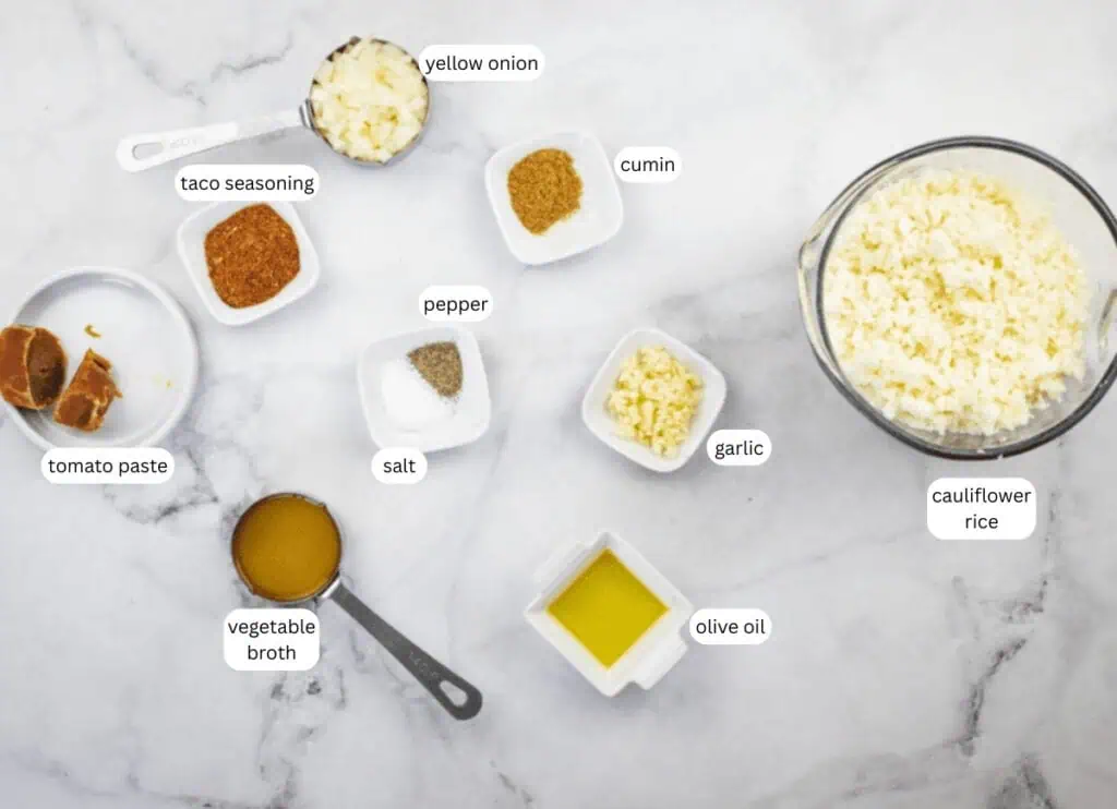 Various ingredients arranged on a marble surface, including cauliflower rice, chopped yellow onion, garlic, cumin, taco seasoning, tomato paste, vegetable broth, olive oil, salt, and pepper.