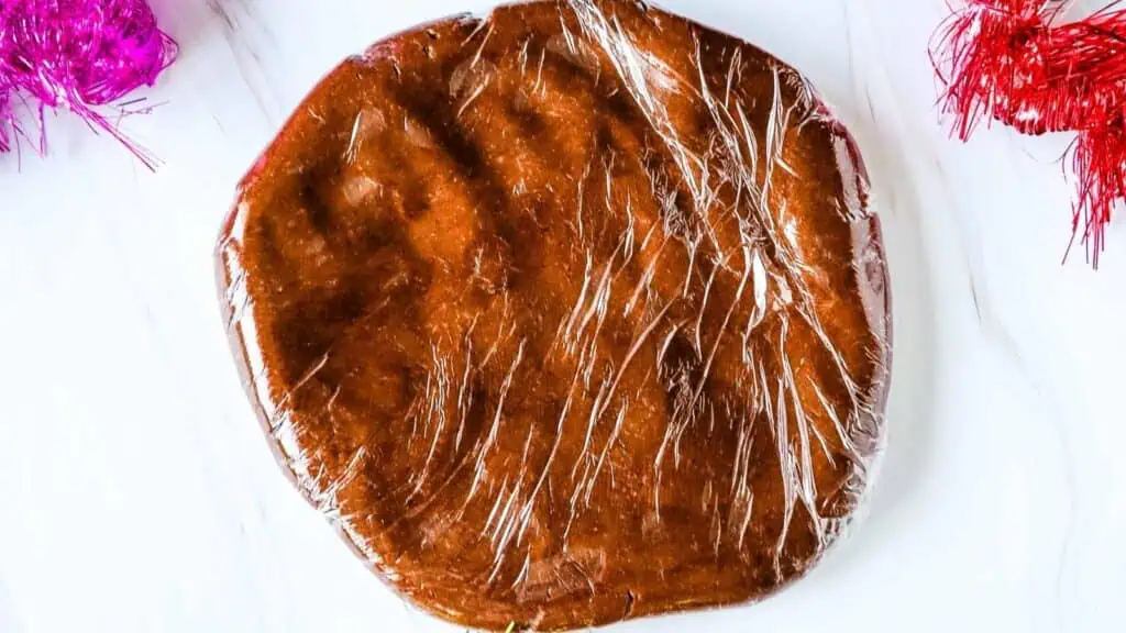 A round disc of chocolate dough wrapped in plastic wrap sits on a white surface with red and pink decorative accents nearby.
