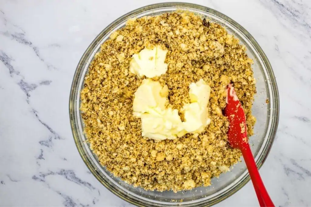 A glass bowl with a crumbly mixture and several chunks of butter, with a red spatula resting inside on a marble surface.