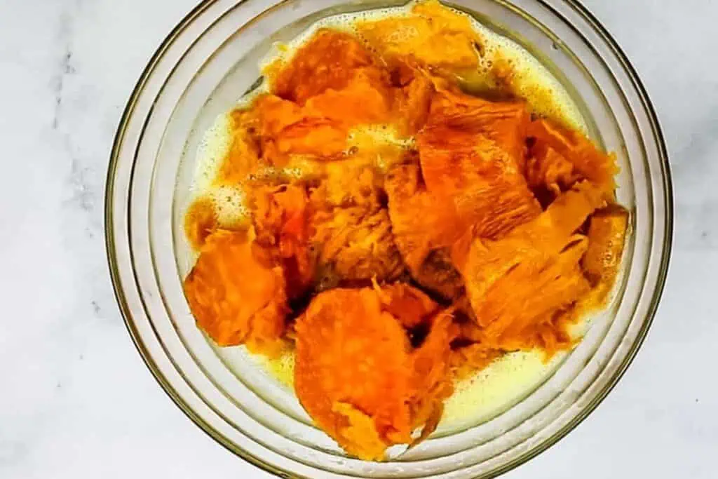 A glass bowl containing mashed cooked sweet potatoes mixed with beaten eggs on a white surface.
