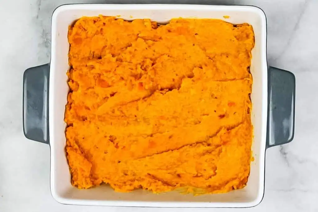 Square white baking dish with grey handles containing an even layer of mashed sweet potatoes, spread smoothly across the surface.