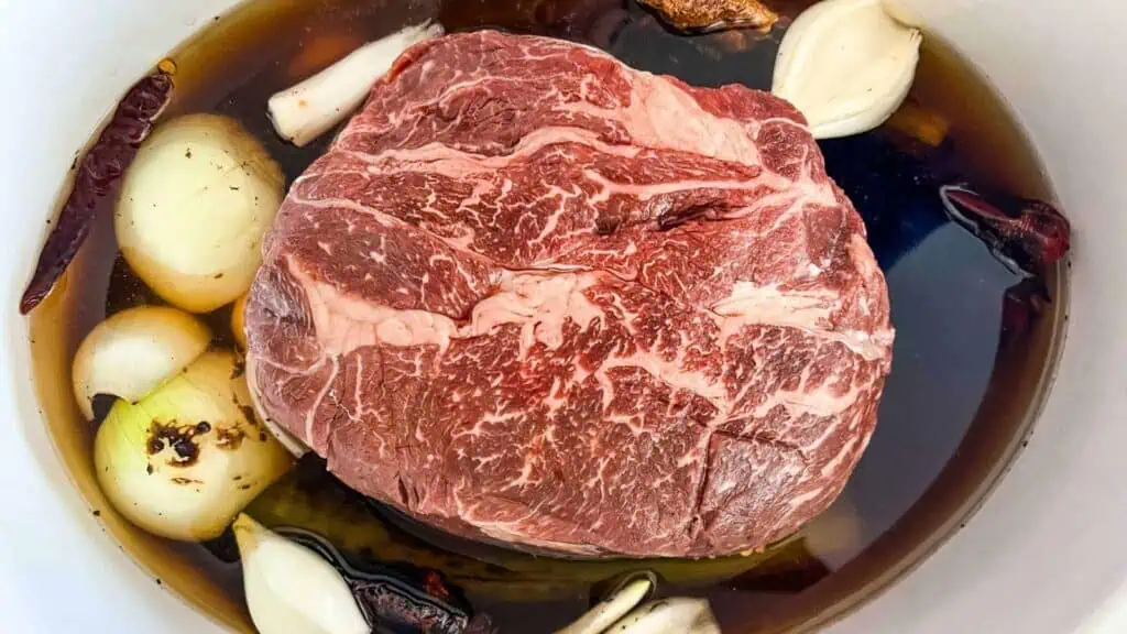 Raw marbled beef roast in a pot with whole garlic cloves, onions, dried chilies, and dark liquid marinade.