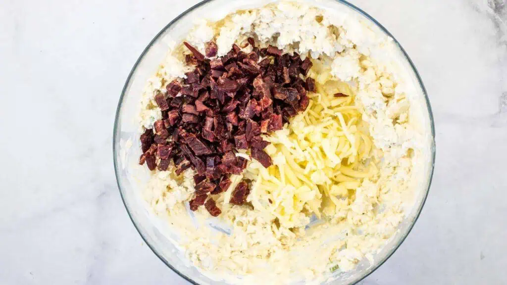 A glass bowl containing a mixture of creamy ingredients, shredded cheese, and chopped dried meat on a light surface.