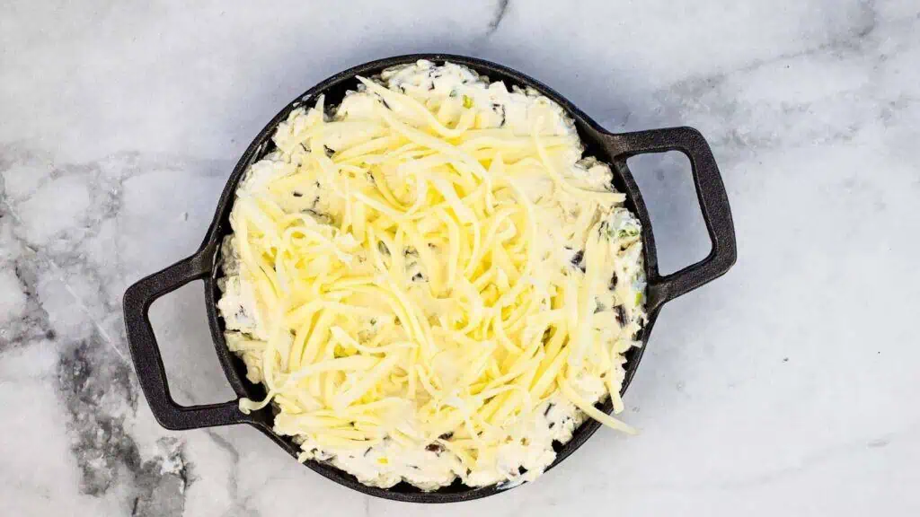 A round black skillet filled with a creamy mixture topped with shredded cheese, placed on a light gray marble surface.