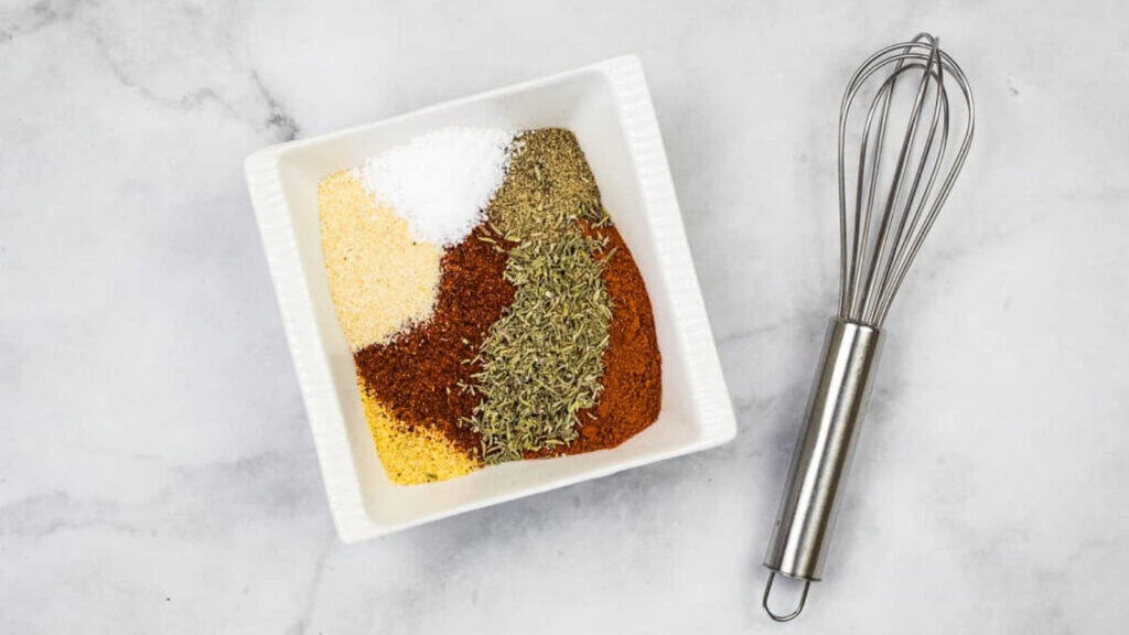A square white bowl with six different spices arranged separately, next to a metal whisk on a light marble surface.